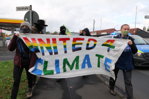 Eine Gruppe maskierter Personen mit einem 'United Climate'-Schild steht vor einer Tankstelle, mit Fahrzeugen auf der Straße, Gras, Schildern, Gebäuden, Bäumen und einem bewölkten Himmel im Hintergrund.