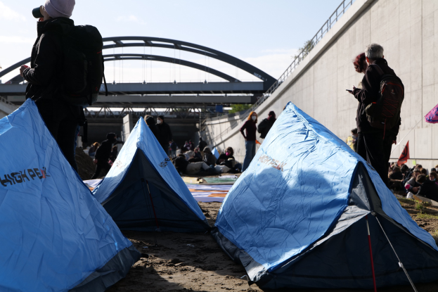 Eine Gruppe von Menschen sitzt auf einem sandigen Strand in der Nähe von Zelten, mit einer Wand und einer Brücke im Hintergrund, die an einer Klimademonstration teilnehmen unter einem bewölkten Himmel.