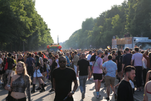 Eine große Menge Menschen, die eine von Bäumen gesäumte Straße entlanggehen, mit einem Turm im Hintergrund und Fahrzeugen mit Menschen auf der rechten Seite, wahrscheinlich beim Christopher Street Day in Berlin unter einem klaren blauen Himmel.
