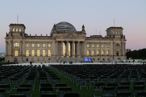 Das Reichstagsgebäude in Berlin, Deutschland, mit seiner markanten Kuppel, Säulen, Bögen und Fenstern, umgeben von Menschen auf einer grünen Fläche mit Stühlen, Straßenlaternen, Schildern, Bäumen und einem bewölkten Himmel.