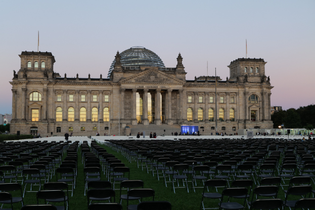 Das Reichstagsgebäude in Berlin, Deutschland, mit seiner markanten Kuppel, Säulen, Bögen und Fenstern, umgeben von Menschen auf einer grünen Fläche mit Stühlen, Straßenlaternen, Schildern, Bäumen und einem bewölkten Himmel.