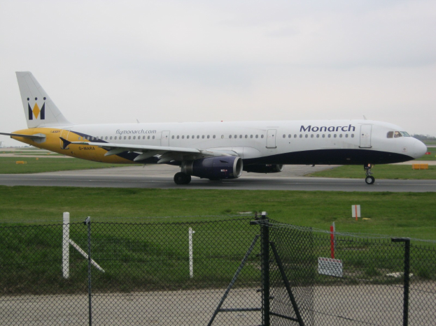 Ein Monarch Airlines Airbus A320-200 auf einer von Gras, einem Zaun mit Pfählen und Brettern, Bäumen und einem klaren blauen Himmel umgebenen Rollbahn.