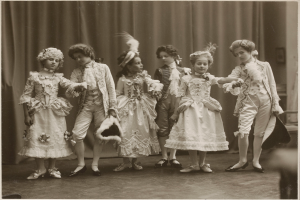 Eine Gruppe von Kindern in Kleidern und Schuhen steht zusammen auf einer Bühne mit einem Vorhang im Hintergrund.