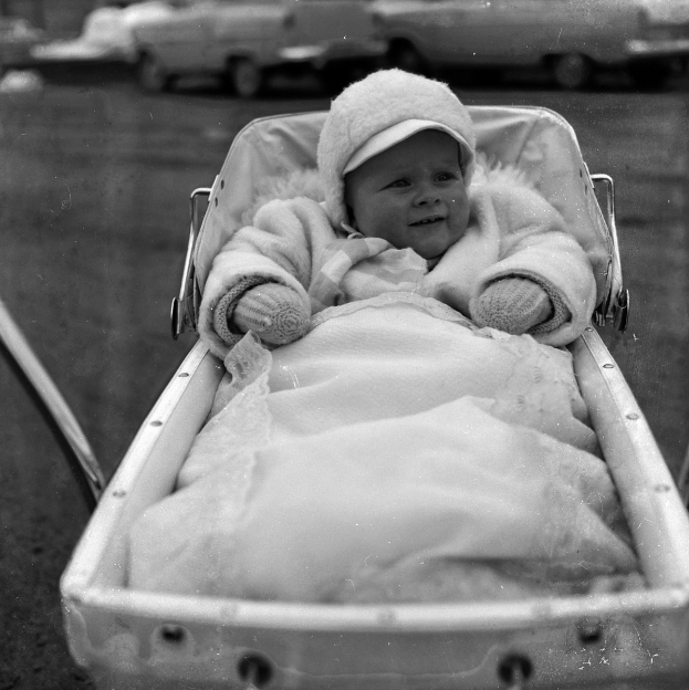 Ein Schwarz-Weiß-Foto eines Babys in einem Kinderwagen, das eine weiße Kleidung und eine Kappe trägt und lacht, mit Fahrzeugen im Hintergrund.