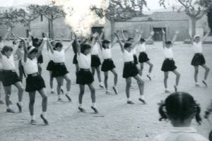 Schwarzes und weißes Foto von jungen Mädchen in Schuluniformen, die auf einer Straße tanzen, die von Gebäuden und Bäumen gesäumt ist, mit einer Person in Schuhen unten.