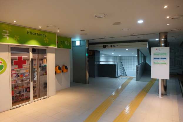 Apothekeneingang mit einem 'Apotheke'-Schild auf der linken Seite, einer Tafel mit Text auf der rechten Seite und beleuchteten Lampen über dem Dach.