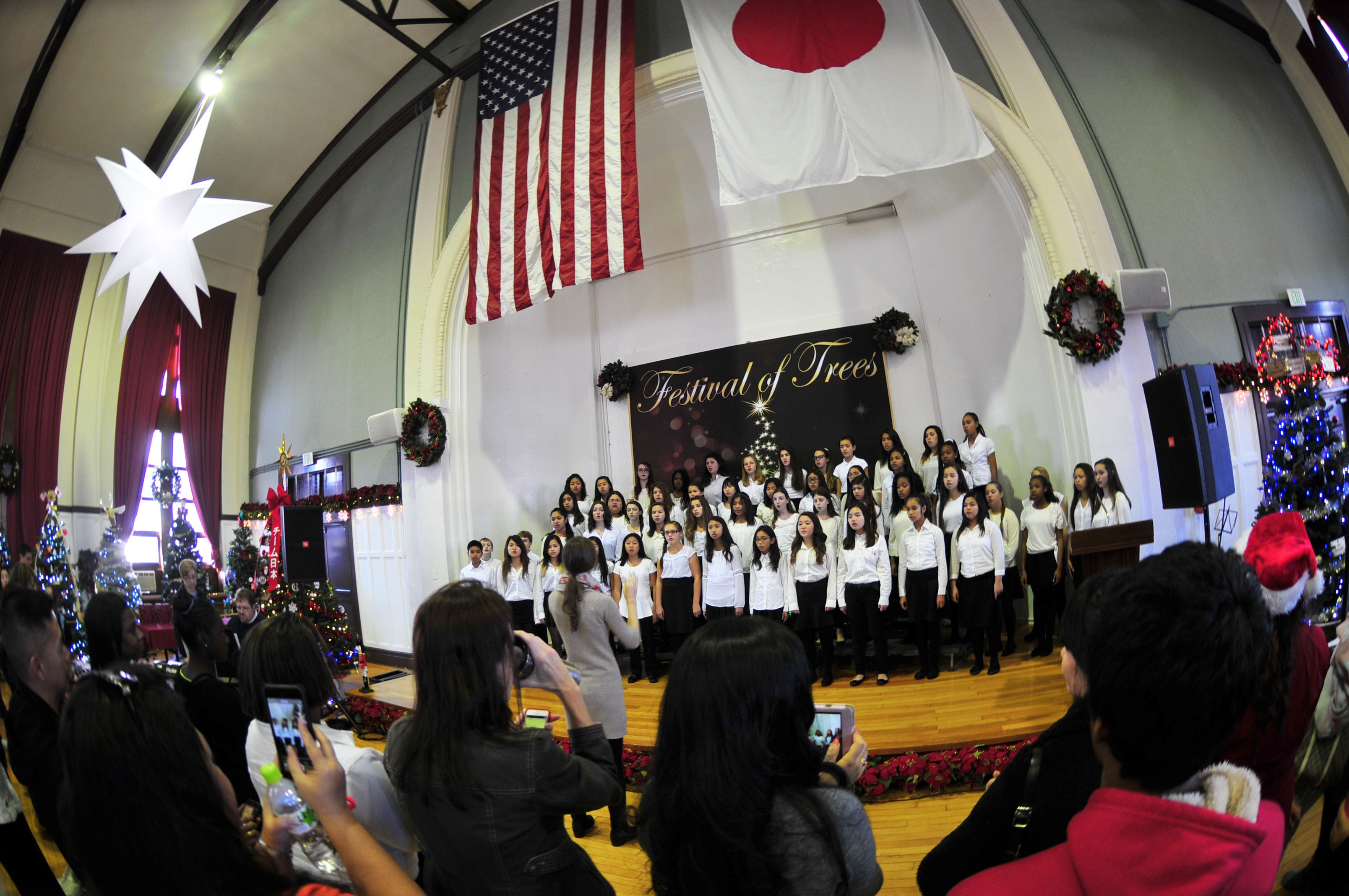 Eine Gruppe von Menschen auf einer Bühne vor einem Publikum, einige halten Kameras und Handys, mit geschmückten Weihnachtsbäumen, einem Podium mit Mikrofon, Lautsprechern, einer Texttafel, Vorhängen, Flaggen und Deckenleuchten.