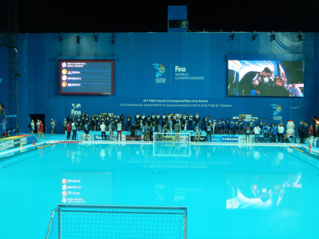 Eine Gruppe von Menschen um ein Hallenbad mit einem Netz am Boden herumstehend, mit einer Wand im Hintergrund, die "FINA World Championships 2015 Kazan" anzeigt.