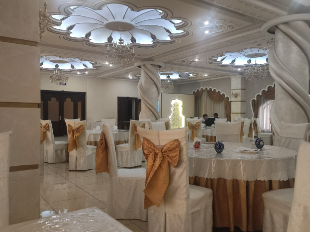 Ein großer Festsaal mit Tischen und Stühlen, die mit Tüchern drapiert und mit verschiedenen Gegenständen dekoriert sind, mit Türen und Vorhängen an den Wänden und beleuchtet von Deckenlampen und Kronleuchtern.