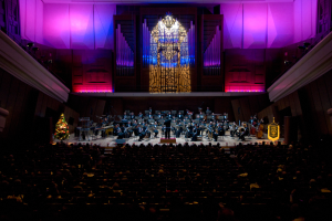 Ein großer Saal voller Menschen, die ein Konzert ansehen, Musiker auf der Bühne, ein Weihnachtsbaum im Hintergrund und Wände mit Lichtern und anderen Gegenständen geschmückt.