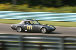 Ein Auto mit einem Helm tragenden Fahrer fährt auf einer Straße, die von Barrieren gesäumt ist, mit Bäumen im Hintergrund.