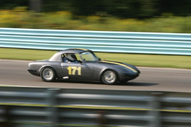 Ein Auto mit einem Helm tragenden Fahrer fährt auf einer Straße, die von Barrieren gesäumt ist, mit Bäumen im Hintergrund.