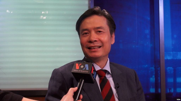 Ein Mann in Anzug und Krawatte hält ein Mikrofon, mit der Hand eines Reporters links daneben, während er in einem Interview vor einem Fenster spricht.