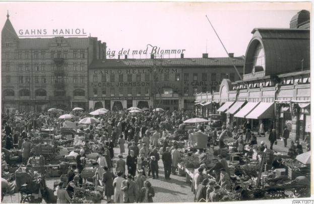 Schwarz-weiß-Foto eines belebten Berliner Markts mit Menschen, Gemüsewagen und Gebäuden im Hintergrund.