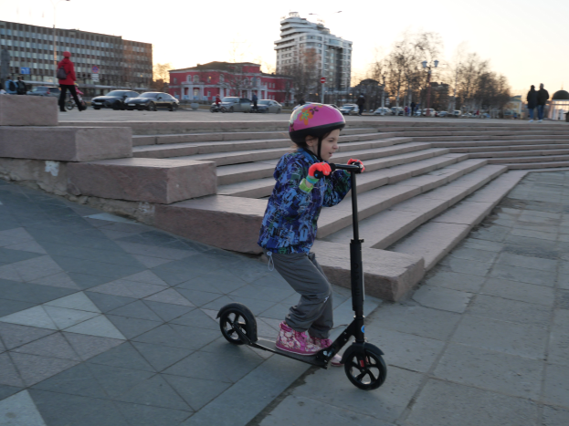 Ein junges Mädchen mit Helm fährt auf einem Gehweg mit Treppen, Fahrzeugen, Menschen, Laternen, Bäumen, Gebäuden und einem klaren blauen Himmel im Hintergrund.