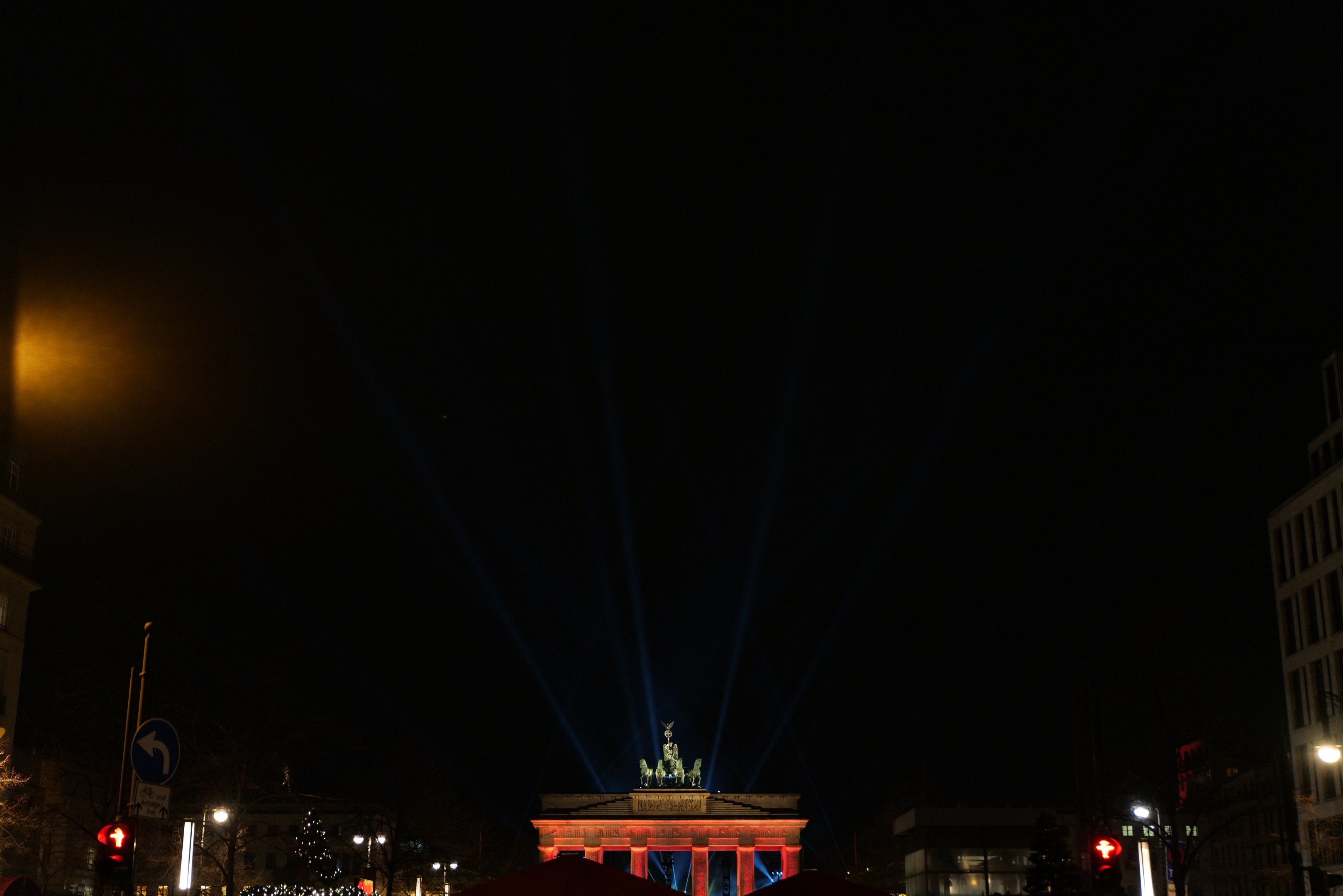 Brandenburger Tor bei Nacht, beleuchtet von Straßenlaternen, mit umliegenden Gebäuden, Bäumen, Pfählen, Verkehrszeichen, Schildern und Zelten gegen einen dunklen Himmel.