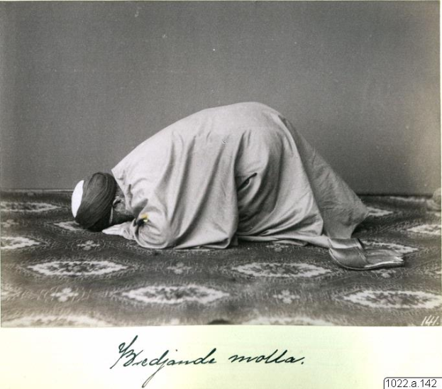 Ein altes Schwarz-Weiß-Foto eines Mannes im Gebet, der ein Tuch und eine Mütze trägt, mit der Beschriftung "Bredjande Molla, die erste Muslimin der Welt" unten.