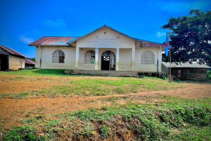 Ein kleines weißes Gebäude mit einer Flagge oben drauf, umgeben von Gras und Bäumen, unter einem klaren blauen Himmel, mit Fenstern, Türen und Text an der Wand, der darauf hinweist, dass es eine Schule für Kinder ist.