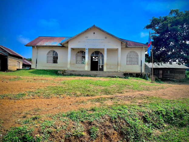 Ein kleines weißes Gebäude mit einer Flagge oben drauf, umgeben von Gras und Bäumen, unter einem klaren blauen Himmel, mit Fenstern, Türen und Text an der Wand, der darauf hinweist, dass es eine Schule für Kinder ist.