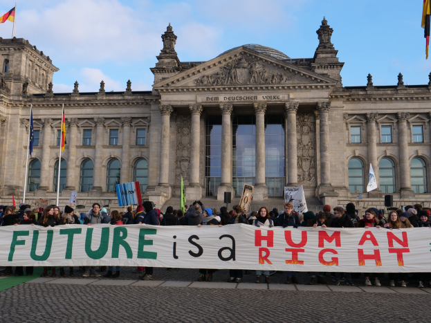 Gruppe von Menschen mit einem Banner mit der Aufschrift "Zukunft ist ein Menschenrecht" vor dem Reichstagsgebäude in Berlin, Deutschland, mit sichtbaren architektonischen Details des Gebäudes und Flaggen im Hintergrund bei bewölktem Himmel.