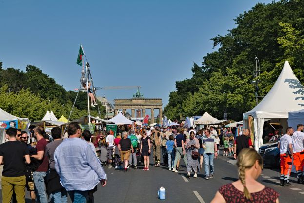 Eine Menschenmenge, die eine Straße mit Zelten, Fahrzeugen und Bäumen entlanggeht, mit einem Bogen im Hintergrund unter einem klaren blauen Himmel und Fahnenmasten auf der linken Seite.
