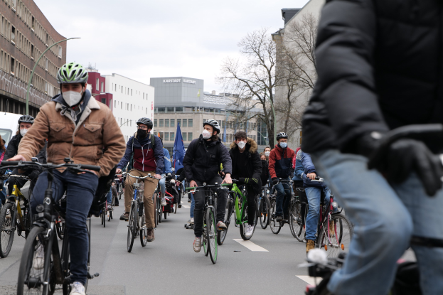 Eine Gruppe von Menschen in Helmen und Handschuhen, die Fahrräder auf einer von Bäumen gesäumten Straße in Berlin fahren, mit Gebäuden, geparkten Fahrzeugen und Himmel im Hintergrund.
