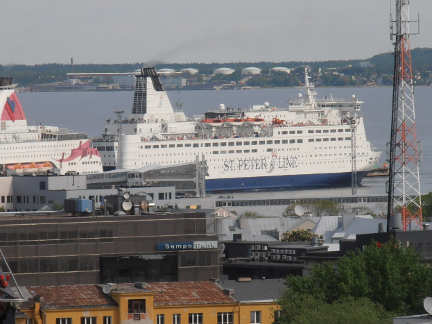 Ein großes Kreuzfahrtschiff liegt in einem Hafen in der Nähe einer Stadt, umgeben von Gebäuden, Bäumen, Türmen und anderen Gegenständen, mit Bäumen, Gebäuden, Hügeln und einem klaren blauen Himmel im Hintergrund.