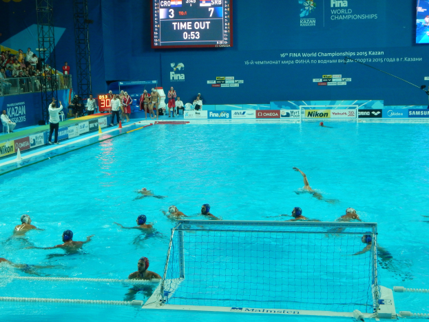 Eine Gruppe von Menschen spielt Wasserball in einem Pool während der 2015 FINA World Championships in Kazan, mit einem Netz, Zuschauern, Tafeln mit Text und Logos, Trägern und einem Bildschirm mit Zahlen und Text im Hintergrund.