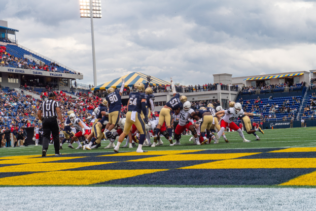 Ein Fußballspiel im Gange mit Spielern in Helmen im Vordergrund, Zuschauern, Geländern, Tafeln mit Text, Laternen und einem bewölkten Himmel im Hintergrund und Text unten auf dem Bild.