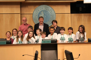 Eine Gruppe von Kindern und Erwachsenen steht vor einem Rednerpult, lacht und posiert für ein Foto, mit Monitoren, Mikrofonen und anderen Gegenständen auf einem Tisch im Vordergrund, einem Logo an der Wand und einem Bildschirm auf der rechten Seite im Hintergrund, bei einer Stadtratssitzung.