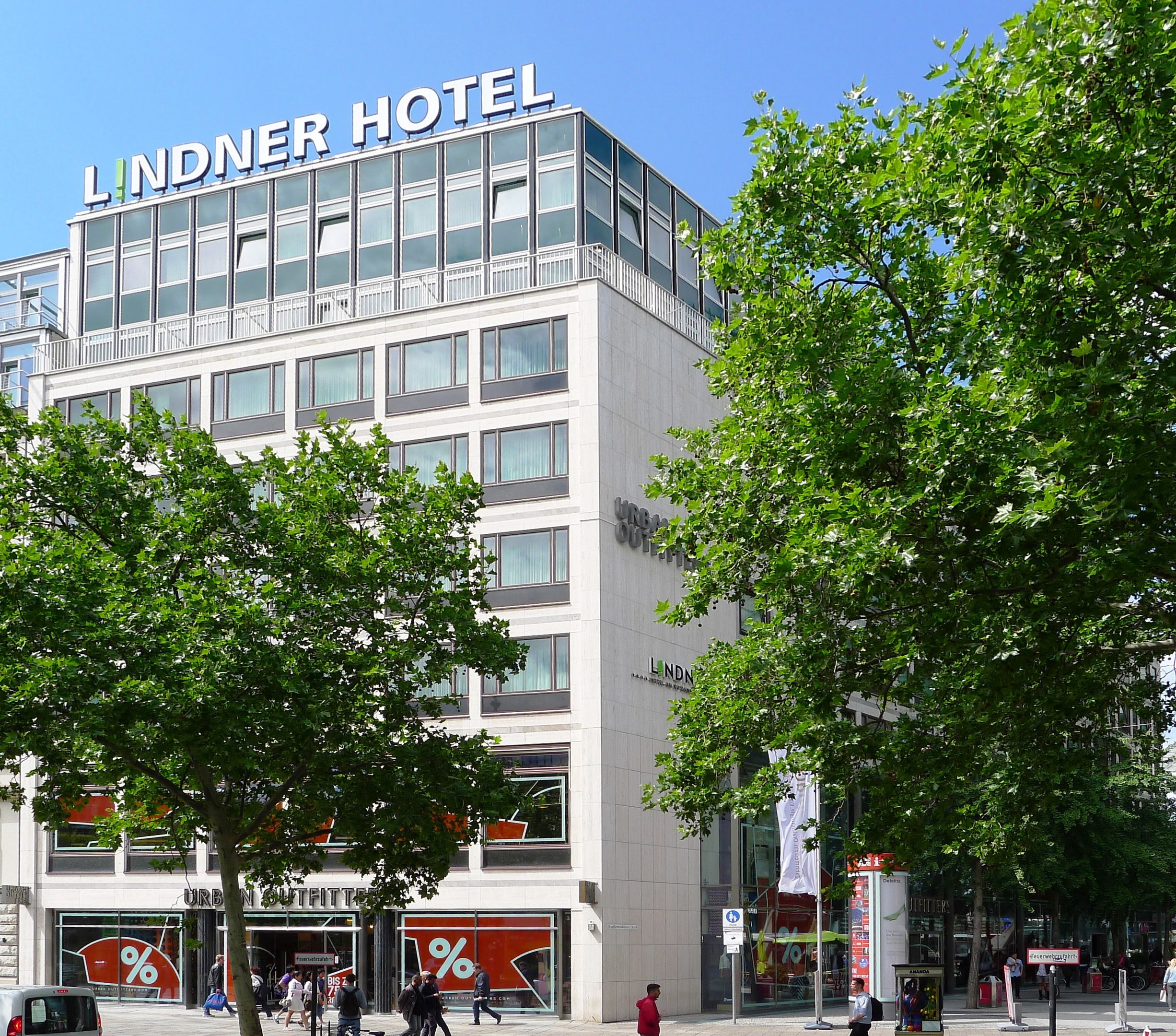 Großes Gebäude mit Glasfront und "Lindner Hotel"-Schriftzug oben, umgeben von Bäumen und einer belebten Straße mit Fußgängern und Fahrzeugen, unter einem sichtbaren Himmel.