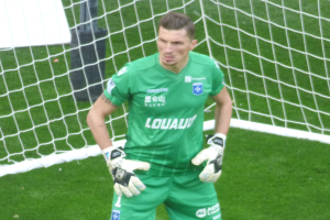 Ein Torwart in grüner Uniform steht vor einem Fußballtor, trägt Handschuhe auf einem Rasenplatz.