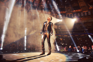 Ein Mann steht auf einer Bühne und hält ein Mikrofon in der Hand, umgeben von Lautsprechern, mit einer Menschenmenge im Hintergrund in einem Stadion, einige halten Kameras, unter hellen Scheinwerfern und einer sichtbaren Decke.