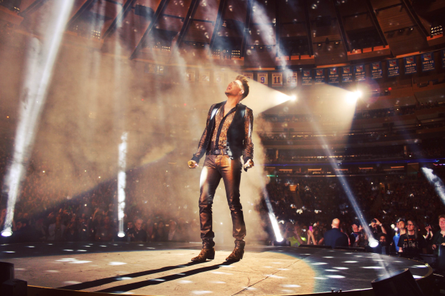 Ein Mann steht auf einer Bühne und hält ein Mikrofon in der Hand, umgeben von Lautsprechern, mit einer Menschenmenge im Hintergrund in einem Stadion, einige halten Kameras, unter hellen Scheinwerfern und einer sichtbaren Decke.