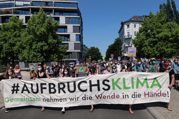Gruppe von Menschen mit Masken, die ein Transparent mit der Aufschrift 'Aufbruchsklima' vor einem Gebäude mit Bäumen und einem klaren Himmel halten.