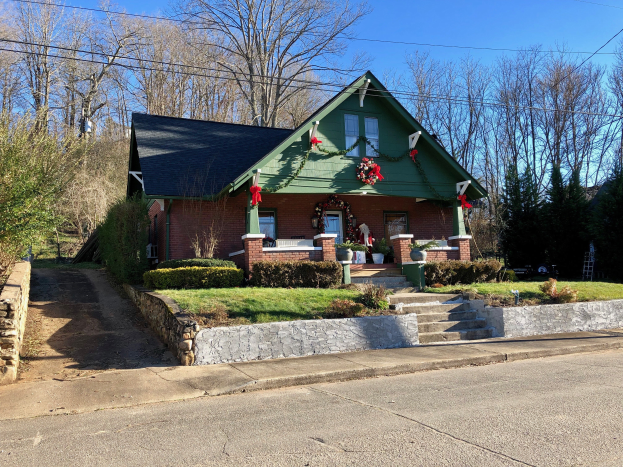 Ein gr├╝nes Haus mit einem Kranz an der Vorderseite, umgeben von Pflanzen, Gras, Treppen, einer Wand, einer Gruppe von B├Ąumen, einigen Dr├Ąhten und einem bew├Âlktem Himmel, gelegen im Herzen der Stadt und zur Miete verf├╝gbar.