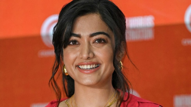Eine Frau in einem roten Kleid mit einem eleganten Dutt, schwarzen High Heels und dezentem Make-up lächelt in die Kamera vor einem gelben Banner mit der Aufschrift "Sonam Kapoors schönste Looks vom Cannes Film Festival."