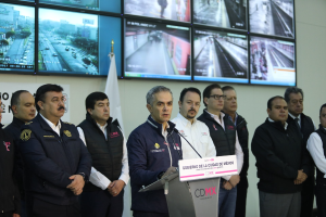 Gruppe von Männern, die vor einem Rednerpult mit einem Mikrofon und einer Tafel mit der Aufschrift "Mexikanische Zoll- und Grenzschutzbehörde" stehen, mit Fernsehern und einer Flagge an der Wand dahinter.