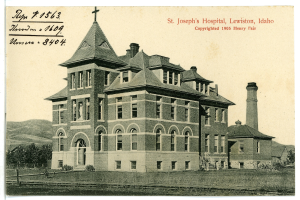 Ein Schwarz-Weiß-Foto des St. Joseph-Krankenhauses in Lewiston, Idaho, das ein großes Gebäude mit Fenstern, eine Treppe mit Geländern, einen Schornstein auf der rechten Seite und Text oben zeigt, mit Bäumen, Hügeln und Himmel im Hintergrund.