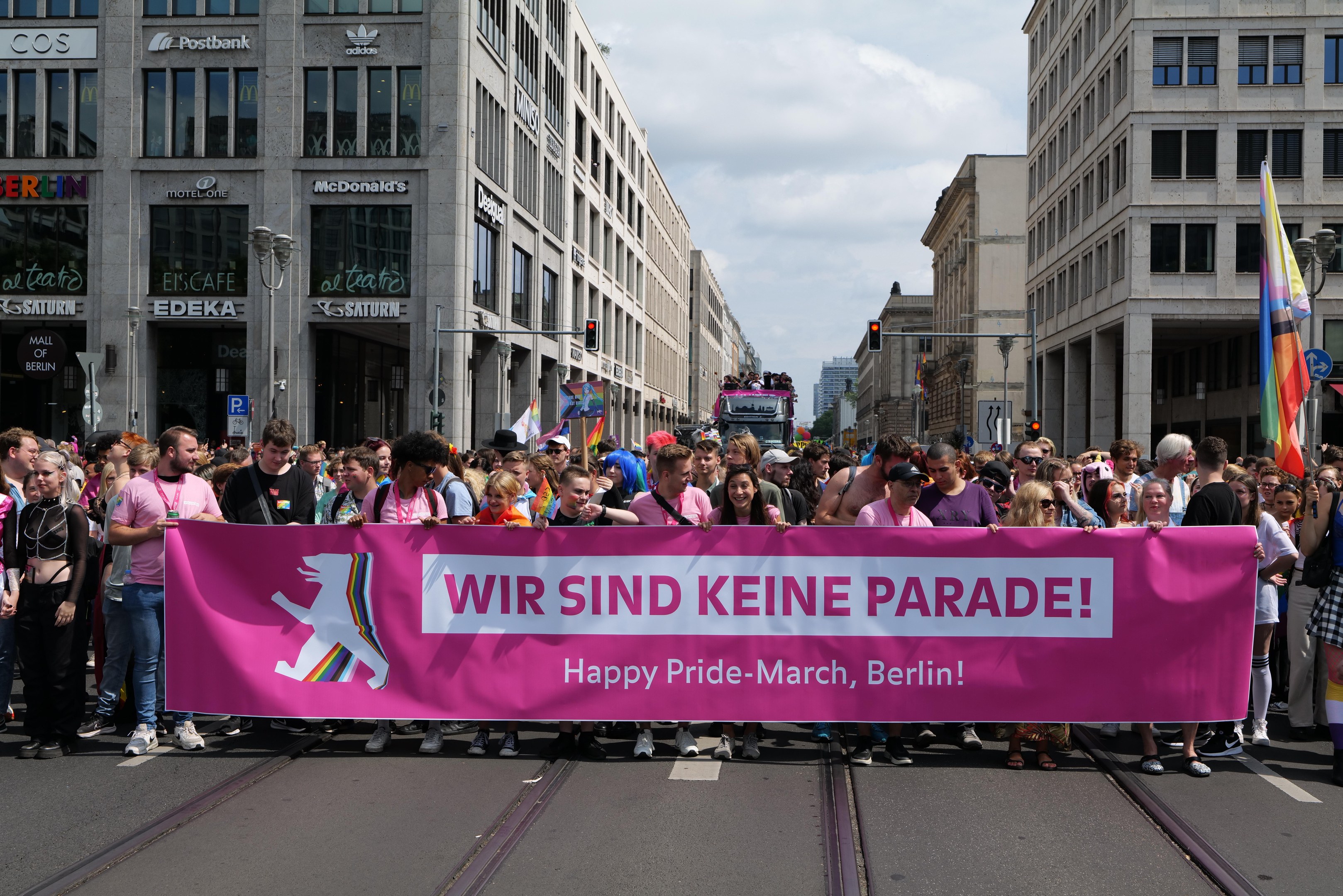 Eine Gruppe von Menschen geht eine Straße in Berlin entlang und hält ein pinkes "Happy Pride March"-Schild, mit Gebäuden, Laternenmasten und Verkehrszeichen an der Straße unter einem bewölkten Himmel.