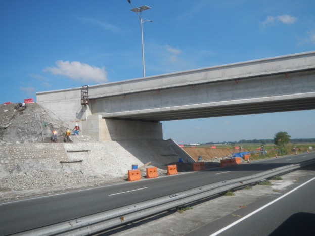 Autobahn mit einer Brücke darüber, ein darunter fahrendes Auto und Bauarbeiter am Straßenrand, markiert durch Verkehrskegel, Steine und einen Laternenmast, mit Bäumen und einem bewölkten Himmel im Hintergrund.