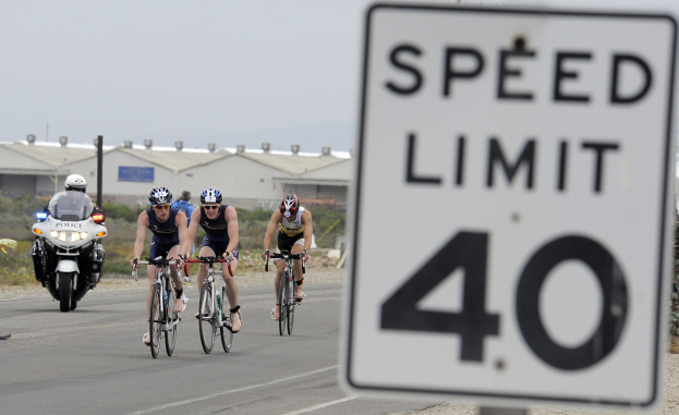 Eine Gruppe von Radfahrern mit Helmen fährt auf einer Straße mit einem Geschwindigkeitsbegrenzungsschild, Gebäuden, Bäumen und einem klaren Himmel im Hintergrund.