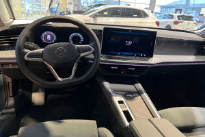 Das Innere eines Volkswagen e-Golf, mit einem Lenkrad, Armaturenbrett, Bildschirmen, Knöpfen, Sitzen und Glastüren, mit Blick auf andere Autos, Gebäude, Bäume, Pfosten und den Himmel durch die Glastüren.