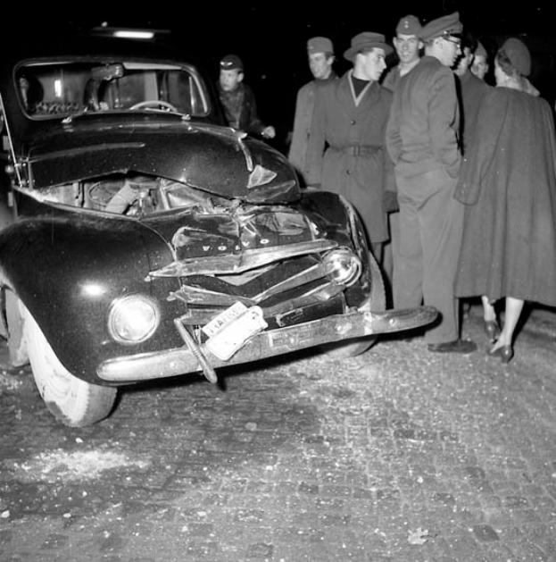 Eine Schwarz-Weiß-Szene eines beschädigten Autos am Straßenrand, mit einer Gruppe von Menschen drumherum, die das Auto besorgt anschauen.