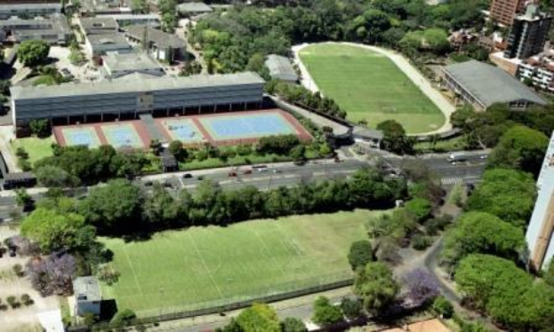 Luftaufnahme eines Fußballfelds an der Universität Sydney, umgeben von Bäumen, Gebäuden und Fahrzeugen, mit einem neuen Sportkomplex in Planung.