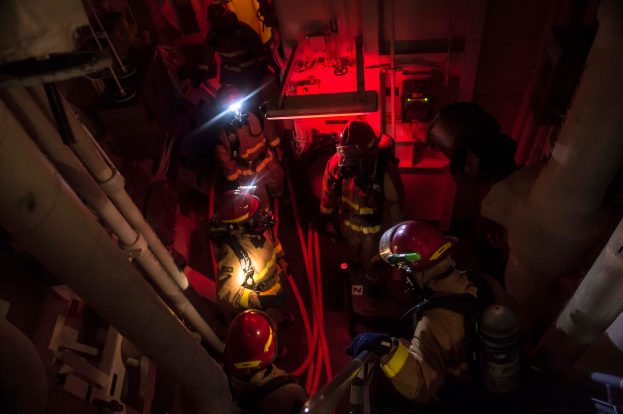Eine Gruppe von Feuerwehrmännern in Helmen und Sauerstoffflaschen steht in einem dunklen Raum mit Rohren auf beiden Seiten, mit einer Maschine und anderen Gegenständen im Hintergrund, was auf einen Rettungseinsatz hinweist.
