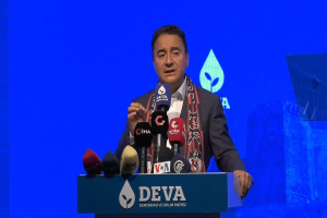 Mann im Mantel und Schal steht hinter Podium mit Mikrofonen und spricht zu Publikum; Podium zeigt Tafel mit Text "deva-Gesch├Ąftsführer sagt, es sei keine gute Idee für die Zukunft", mit Bildschirm und Objekt im Hintergrund.