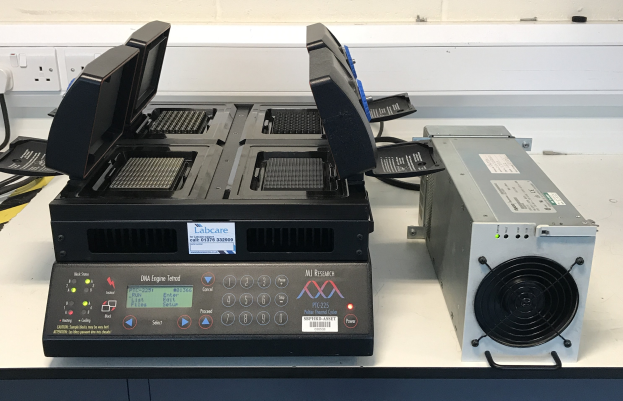 Zwei elektronische Geräte auf einem weißen Tisch in einem Labor, mit einem als DNA-Engine identifizierten und einer Schalttafel an der Wand im Hintergrund.