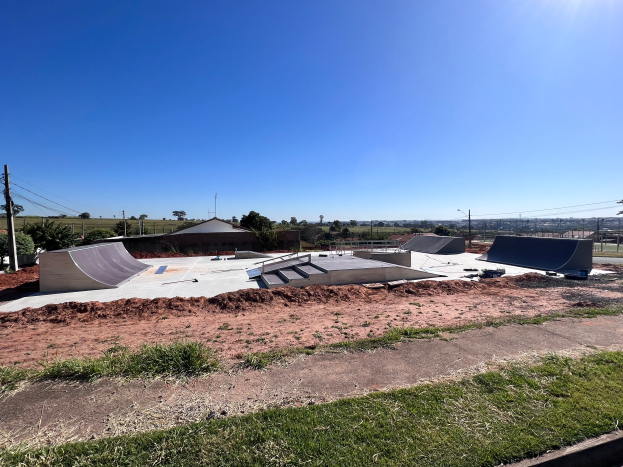 Ein Skatepark mit mehreren Rampen, umgeben von Gras und Bäumen, mit Polen und Drähten im Hintergrund unter einem klaren blauen Himmel.