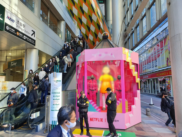 Eine Gruppe von Menschen steht vor einem Gebäude mit Glaswänden und Texttafeln, einige tragen Masken, während eine Rolltreppe mit Fahrgästen zu sehen ist; eine pinkfarbene Box in der Mitte zeigt Schaufensterpuppen und trägt den Text "Netflix's new Netflix-Booth in Tokyo."